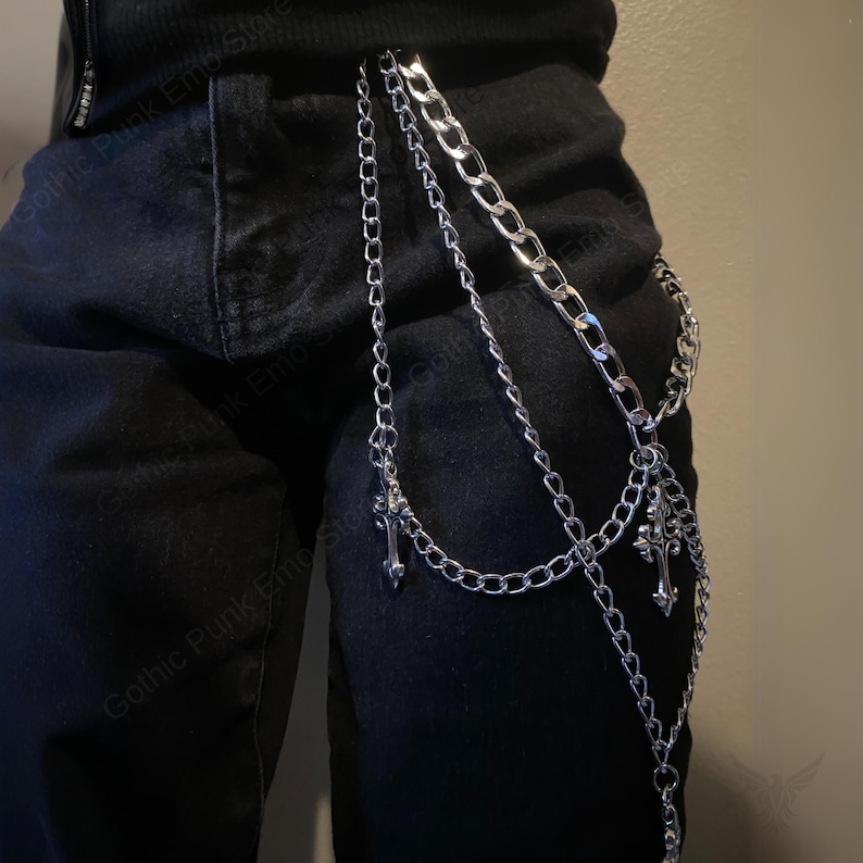 May include: Silver-colored multi-chain belt with cross pendants. The chains are of varying lengths and styles, attached to a black garment. The belt is designed to hang from the waist to the upper thigh.