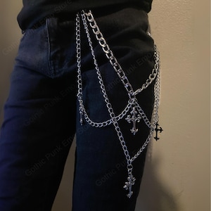 May include: Silver-colored chain belt with multiple strands and decorative cross pendants. The belt is designed to hang from the waist and features various chain styles. The image shows the belt against a dark background, highlighting its metallic shine.