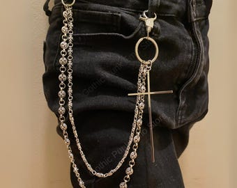 May include: Silver-toned chain accessory with a cross pendant and decorative ball chain. The chain is attached to the belt loops of black denim pants with two metal clasps. The accessory adds a gothic or punk aesthetic.