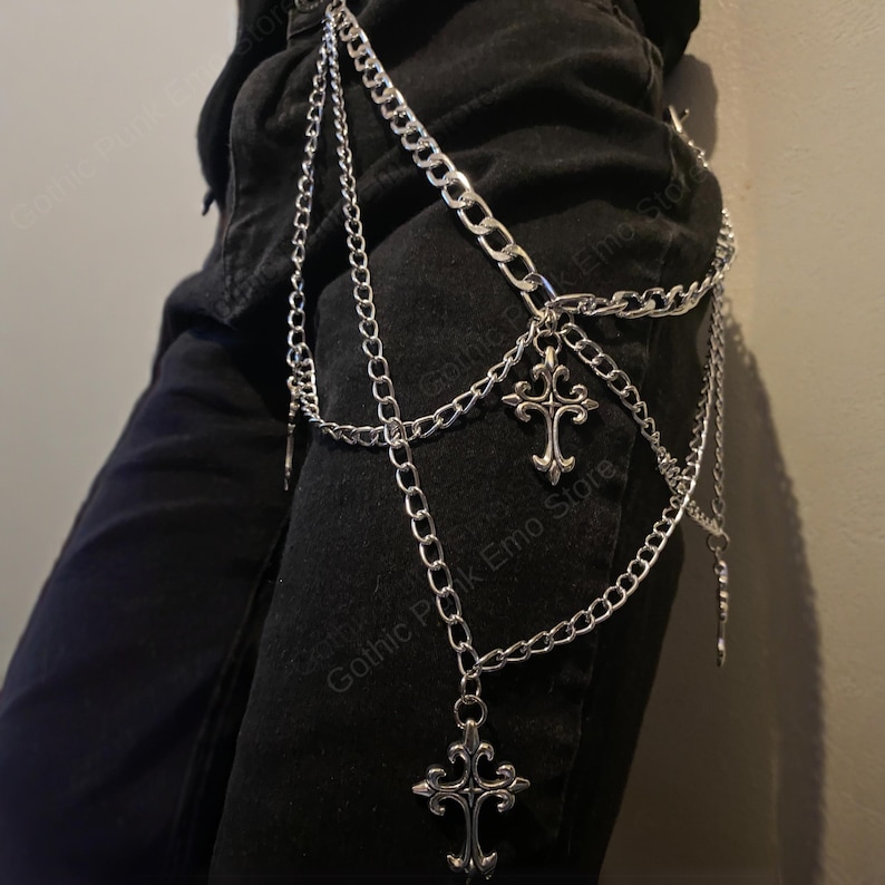 May include: Silver-colored chain accessory with three decorative crosses. The chain is layered and hangs from the waist, creating a gothic or punk aesthetic. The crosses have ornate detailing.