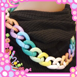 Puede incluir: Un collar de cadena grueso y colorido. El collar presenta eslabones grandes e interconectados en una variedad de colores pastel, incluyendo rosa, morado, azul, verde, amarillo y blanco. El collar se usa sobre un fondo negro.