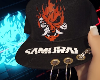May include: Black baseball cap featuring a red and white graphic of a samurai-style demon. The word "SAMURAI" is printed in white on the brim. The brim also has silver hoops and a chain. A stylish accessory.