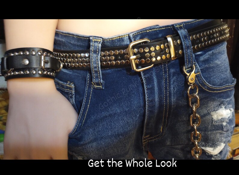 K&ouml;nnte beinhalten: Ein schwarzer Lederg&uuml;rtel mit silbernen Nieten und einer goldenen Schnalle. Der G&uuml;rtel wird &uuml;ber einer blauen Jeans getragen, mit einem passenden schwarzen Lederarmband mit silbernen Nieten. Eine goldene Kette h&auml;ngt an der G&uuml;rtelschlaufe.