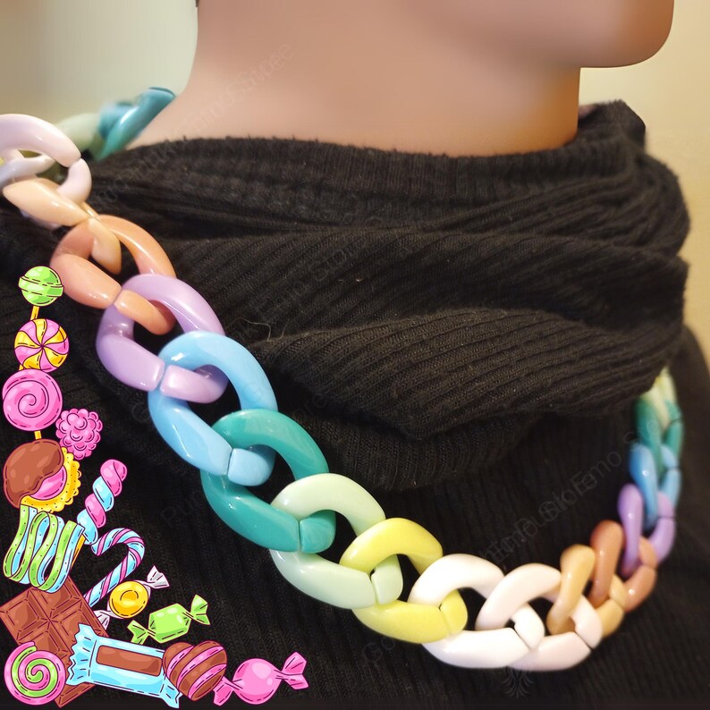 Puede incluir: Un collar de cadena grueso y colorido. El collar presenta eslabones grandes e interconectados en una variedad de colores pastel, incluyendo rosa, azul, verde y amarillo. El collar se usa sobre un fondo oscuro, resaltando los colores vibrantes de la cadena.
