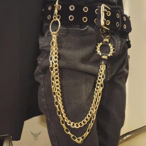 May include: A black belt with gold-colored grommets and a gold buckle. A gold chain hangs from the belt, attached to a gold ring and a decorative ring with spikes. The chain is layered and hangs down the side of the pants.