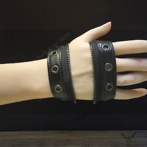 May include: Black leather wrist cuffs with a zipper detail and silver-tone grommets. The cuffs are designed with a punk or gothic aesthetic. The image shows two cuffs, one on each side of the wrist.