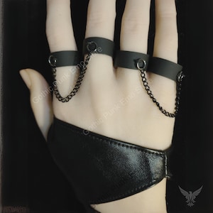May include: Black finger rings connected by chains, with a matching wrist cuff. The rings are made of a matte black material and are connected by delicate black chains. The wrist cuff is made of a textured black material.