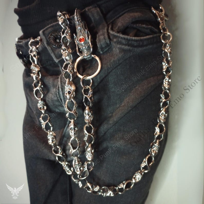 May include: Silver-toned metal wallet chain with skull and dragon head details. The chain is attached to a pair of black jeans. The dragon head has a red gem eye.