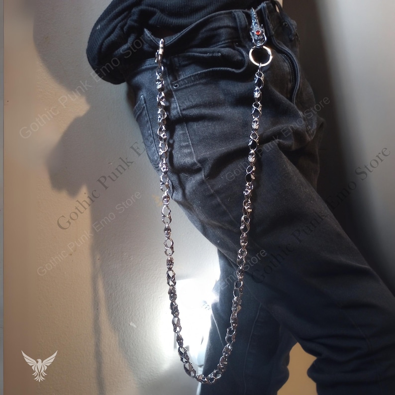 May include: A silver-colored, skull-linked chain, attached to a belt loop and pocket. The chain is made of small, detailed skulls and connects to a larger ring. The image shows the chain hanging down the side of a person's leg.