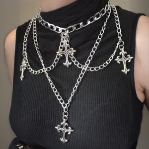May include: Silver-toned layered necklace featuring multiple chains and decorative cross pendants. The necklace has a choker-style design with several strands cascading down. The crosses have ornate detailing, creating a gothic or punk aesthetic.