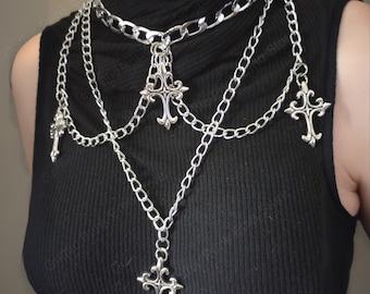 May include: Silver-toned layered necklace featuring multiple chains and decorative cross pendants. The necklace has a choker-style design with several strands cascading down. The crosses have ornate detailing, creating a gothic or punk aesthetic.