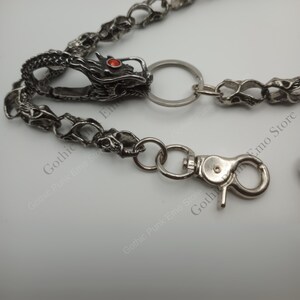 May include: Silver-toned metal wallet chain featuring a dragon head with a red gem eye. The chain has skull-shaped links and a lobster clasp. The dragon head is attached to a key ring.