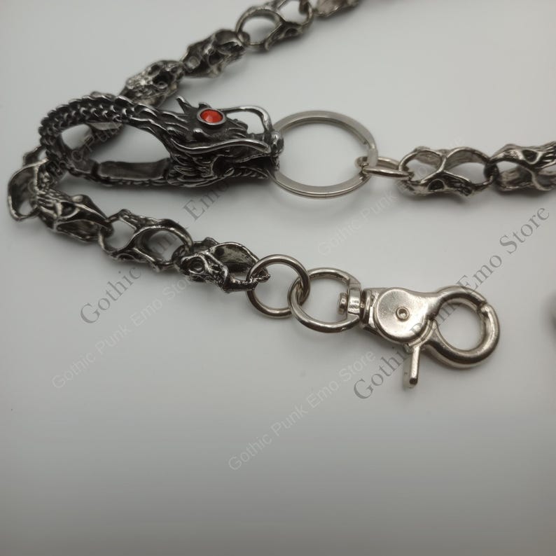 May include: Silver-toned metal wallet chain featuring a dragon head with a red gem eye. The chain has skull-shaped links and a lobster clasp. The dragon head is attached to a key ring.
