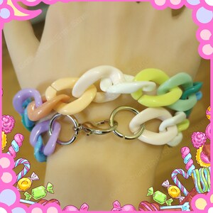 Puede incluir: Una pulsera de cadena colorida con eslabones grandes y redondeados en tonos pastel de rosa, morado, amarillo, verde y blanco. La pulsera se asegura con un cierre y anillos plateados.