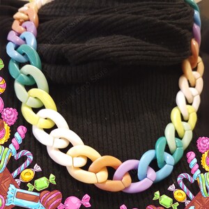Puede incluir: Un collar de cadena gruesa con eslabones ovalados grandes en tonos pastel como rosa, azul, verde, amarillo y blanco. El collar se usa sobre un fondo negro, creando un fuerte contraste. Esta llamativa pieza es un accesorio de moda llamativo.