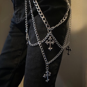 May include: Silver-colored chain leg harness with multiple strands and decorative cross pendants. The harness is designed to be worn on the leg and features a series of interconnected chains. The style is reminiscent of gothic or punk fashion.