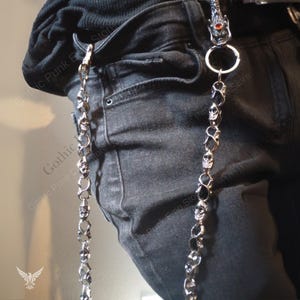 May include: Silver-toned metal chain with skull and black stone accents. The chain is attached to a belt loop and hangs down the side of black denim jeans. The top of the chain features a dragon head with a red gem.