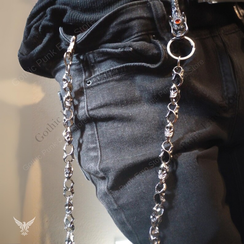 May include: Silver-toned metal chain with skull and black stone accents. The chain is attached to a belt loop and hangs down the side of black denim jeans. The top of the chain features a dragon head with a red gem.