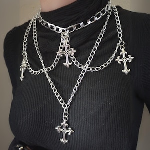 May include: A silver-colored layered necklace with multiple chains and decorative cross pendants. The necklace features a choker-style chain at the top, with cascading chains of varying lengths. The crosses have ornate, gothic-inspired designs.
