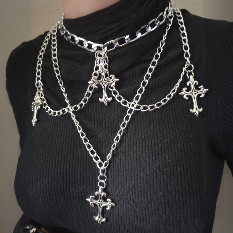 May include: A silver-colored layered necklace with multiple chains and decorative cross pendants. The necklace features a choker-style chain at the top, with cascading chains of varying lengths. The crosses have ornate, gothic-inspired designs.
