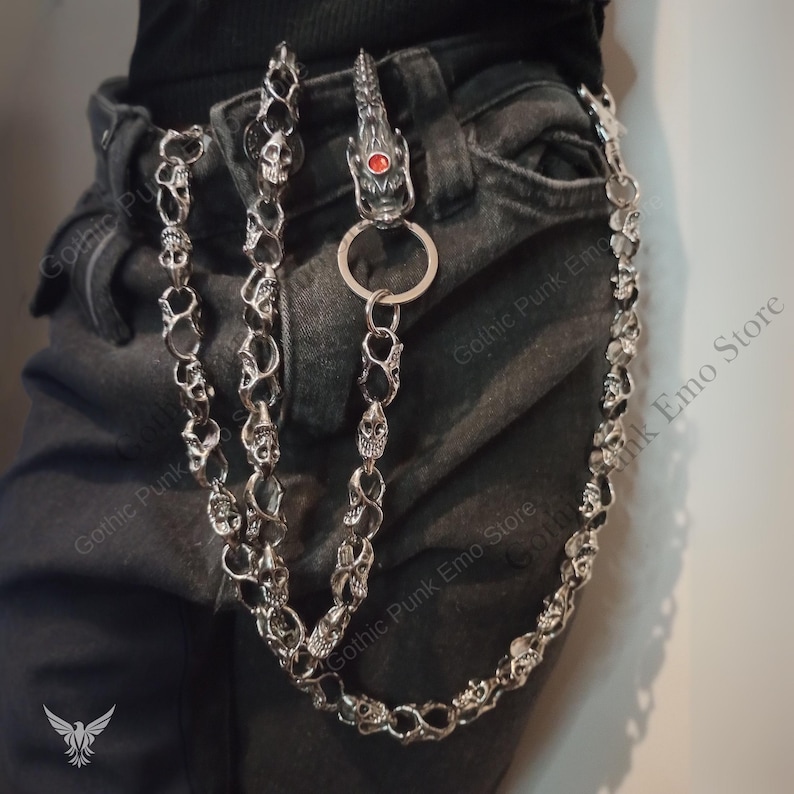 May include: Silver-toned wallet chain featuring skull and dragon head details. The chain is attached to a belt loop, with a dragon head clasp with a red gem. The chain is made of multiple skull links.