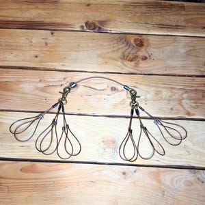May include: A copper-colored wire leash attachment with two brass-colored clasps. The wire is shaped into teardrop loops, creating a decorative and functional design. The attachment is designed to connect to a dog leash.