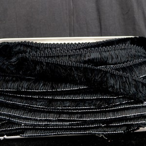 May include: Black fringe trim with a white thread running through it. The trim is folded and stacked on a white surface.