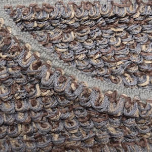 May include: A textured fabric featuring a dense, looped pile construction in shades of grey, brown, and beige. The material displays a repeating pattern of small, raised loops arranged in rows, with a distinct diagonal seam or fold visible.
