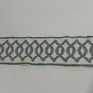 May include: A decorative trim featuring a repeating blue and beige geometric pattern. The trim has a light beige background with a blue interlocking design. A thin blue border runs along the top and bottom edges of the trim.