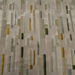 May include: A textile fabric featuring a repeating abstract pattern of vertical rectangular blocks in shades of mustard yellow, olive green, slate blue, and charcoal gray set against a light beige background with a textured, woven appearance.