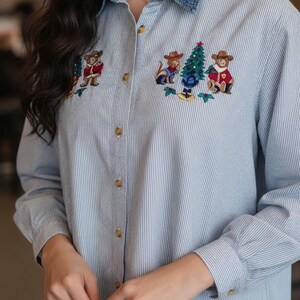 May include: A light blue and white pinstriped button-down shirt featuring a denim collar and embroidered holiday-themed patches. The embroidery depicts two brown bears wearing cowboy hats and red vests flanking a decorated Christmas tree.