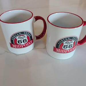 Puede incluir: Dos tazas de cerámica blanca con bordes y asas rojas. Cada taza presenta un gráfico circular con el texto Adrian, Texas, Route US 66 y Midpoint. El diseño incluye un emblema en forma de escudo negro y rojo en la parte frontal de cada taza.
