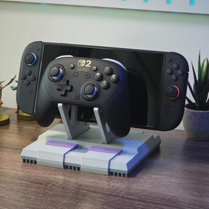 May include: A black Nintendo Switch console and controller rest on a gray and purple stand. The stand is designed to resemble a classic video game console. Small potted plants and figurines are in the background.