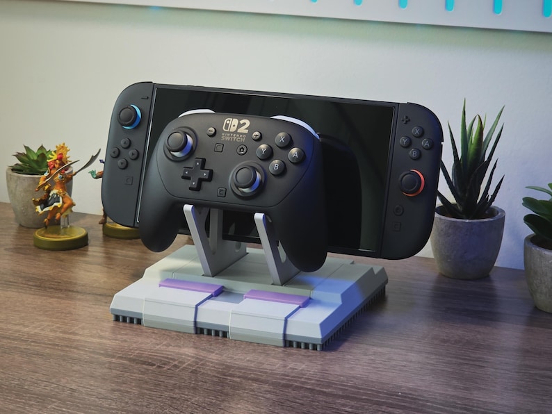 May include: A black Nintendo Switch console and controller rest on a gray and purple stand. The stand is designed to resemble a classic video game console. Small potted plants and figurines are in the background.
