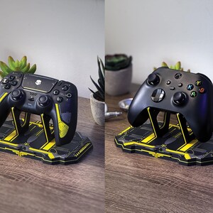 以下が含まれることがあります： 黒と黄色のゲームコントローラーが、お揃いのスタンドに置かれています。 コントローラーにはスカルのロゴと黄色のアクセントがあります。 スタンドは黒で、黄色の線と幾何学模様のデザインが施されています。 コントローラーは木製の表面に置かれています。