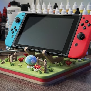 Pode incluir: Uma consola Nintendo Switch com comandos Joy-Con azuis e vermelhos repousa sobre um suporte decorativo. O suporte apresenta uma paisagem verde com um cogumelo azul, flores vermelhas e uma cerca de madeira. O suporte &eacute; de madeira e tem uma borda vermelha.