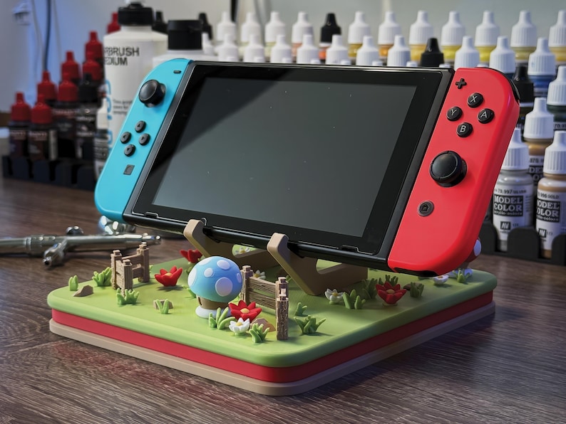 Pode incluir: Uma consola Nintendo Switch com comandos Joy-Con azuis e vermelhos repousa sobre um suporte decorativo. O suporte apresenta uma paisagem verde com um cogumelo azul, flores vermelhas e uma cerca de madeira. O suporte &eacute; de madeira e tem uma borda vermelha.
