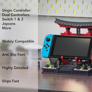 May include: A black, red, and blue Switch console stand with a Japanese Torii gate design. The stand holds the Switch console and Joy-Cons. Text on the side reads "4-in-1", "Widely Compatible", "Anti Slip Feet", and "Highly Detailed".