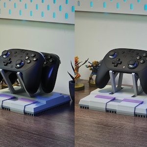 May include: Black game controllers on a gray and purple stand. The stand is designed to resemble a classic video game console. The controllers are supported by a silver-colored frame. The setup is on a wooden surface.