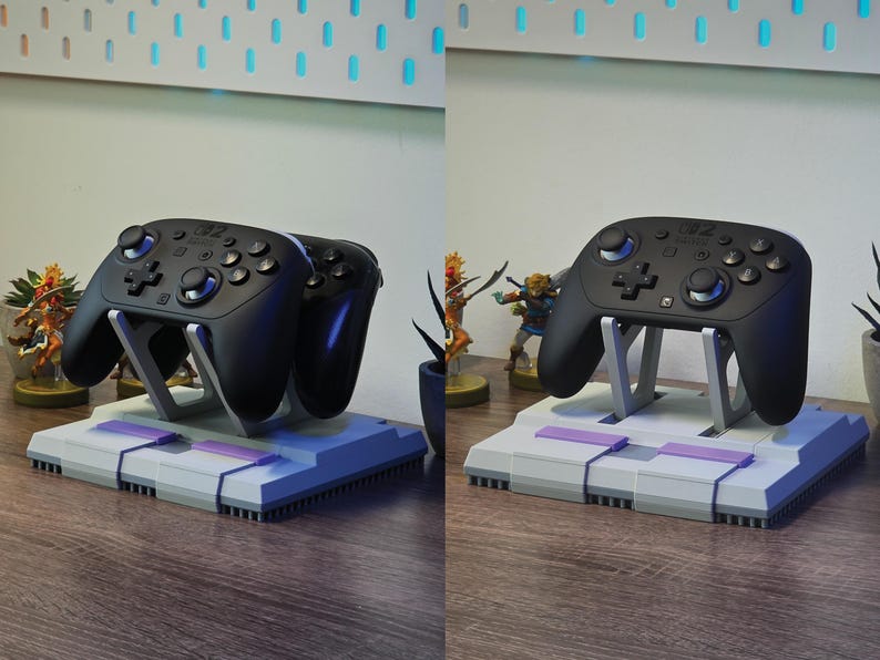 May include: Black game controllers on a gray and purple stand. The stand is designed to resemble a classic video game console. The controllers are supported by a silver-colored frame. The setup is on a wooden surface.