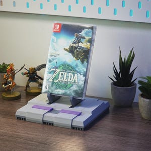 May include: A gray and purple video game console-style stand holding a Nintendo Switch game case. The game case features the title "The Legend of Zelda: Tears of the Kingdom." Small potted plants and figurines are also visible.