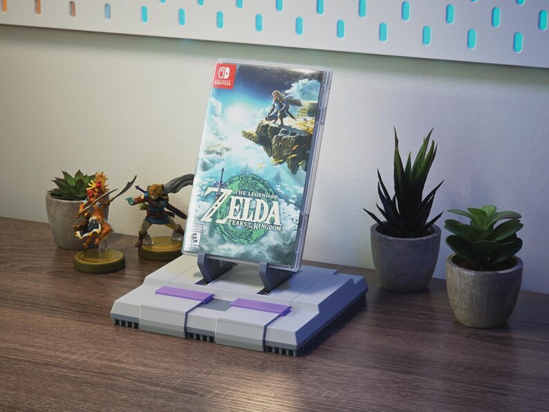 May include: A gray and purple video game console-style stand holding a Nintendo Switch game case. The game case features the title "The Legend of Zelda: Tears of the Kingdom." Small potted plants and figurines are also visible.