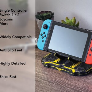以下が含まれることがあります： 黒と黄色のSwitchコンソールスタンド。Switchコンソールが配置されています。スタンドは幾何学的なデザインで、「3-in-1」、「Single Controller」、「Widely Compatible」、「Anti Slip Feet」の文字があります。Switchコンソールには青と赤のコントローラーが付いています。