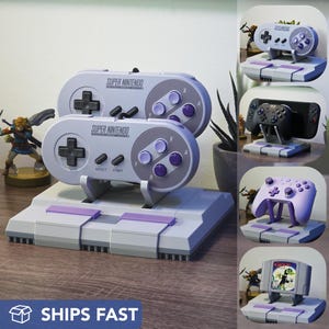 May include: A gray Super Nintendo controller stand with a matching console base. The controller has purple buttons and the text "SUPER NINTENDO". The image also shows other gaming controller stands.