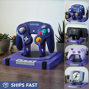 May include: A purple Nintendo GameCube controller on a matching stand. The controller has colourful buttons and the words "Nintendo GameCube" printed on it. The stand has a base with a slot for the controller. The image also shows other game controllers.