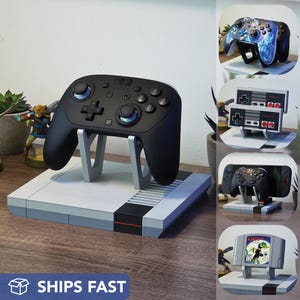May include: A black video game controller rests on a gray and white stand designed to resemble a classic game console. The stand has a black stripe and the text "SHIPS FAST" is visible.