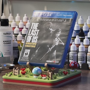Pode incluir: Uma caixa de jogo PS4 para "The Last of Us: Remastered" &eacute; exibida em um suporte decorativo. O suporte apresenta um jardim em miniatura com flores vermelhas, cogumelos e uma cerca de madeira. Frascos de tinta e suprimentos de aer&oacute;grafo ao fundo.