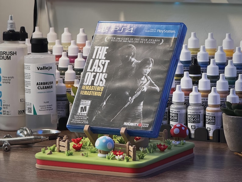 Pode incluir: Uma caixa de jogo PS4 para "The Last of Us: Remastered" &eacute; exibida em um suporte decorativo. O suporte apresenta um jardim em miniatura com flores vermelhas, cogumelos e uma cerca de madeira. Frascos de tinta e suprimentos de aer&oacute;grafo ao fundo.