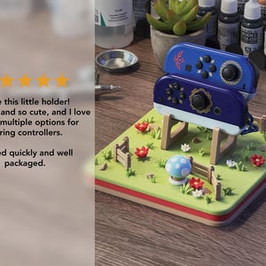 Pode incluir: Um suporte decorativo para controladores com uma base verde e uma borda vermelha e marrom. O suporte tem um design de paisagem com cogumelos, flores e uma cerca. Dois controladores azuis s&atilde;o colocados em cima.