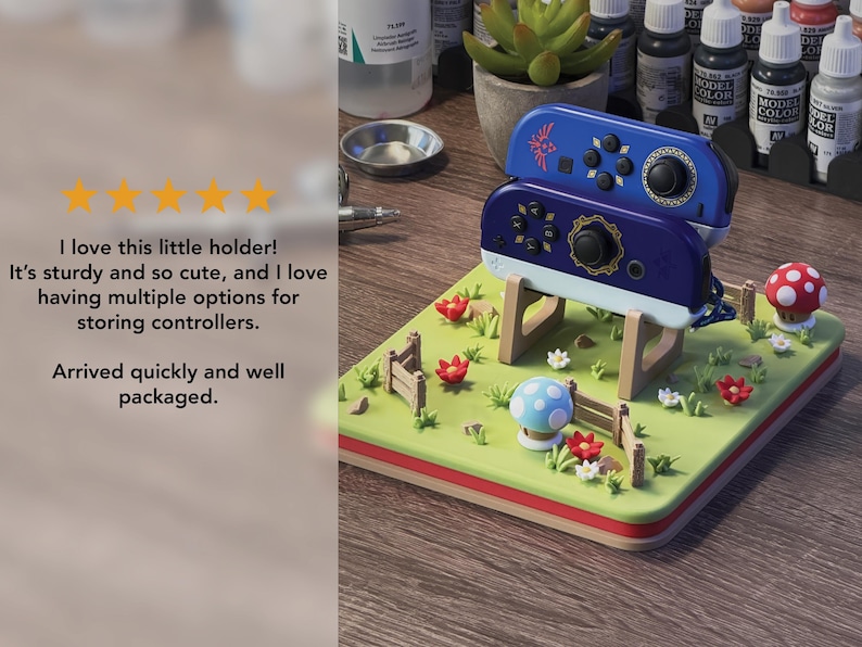 Pode incluir: Um suporte decorativo para controladores com uma base verde e uma borda vermelha e marrom. O suporte tem um design de paisagem com cogumelos, flores e uma cerca. Dois controladores azuis s&atilde;o colocados em cima.